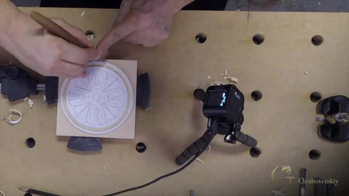 A floral rosette pattern is penciled onto a round wood block clamped in a vise, a small camera set up nearby.