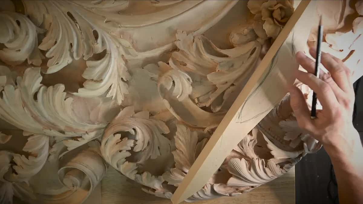 An elaborate carved leaf and scroll panel fills the frame while a hand sketches lines on a wood board laid across it.