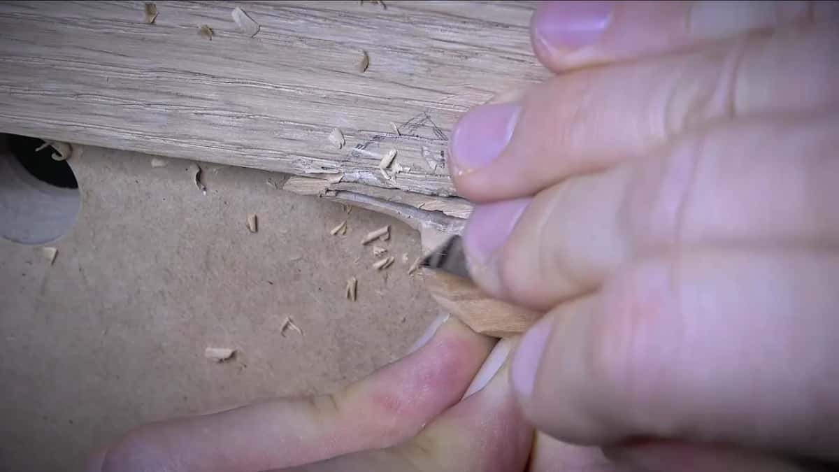 Painted fingernails frame a small blade slicing a thin pale wood sliver near a workbench hole.
