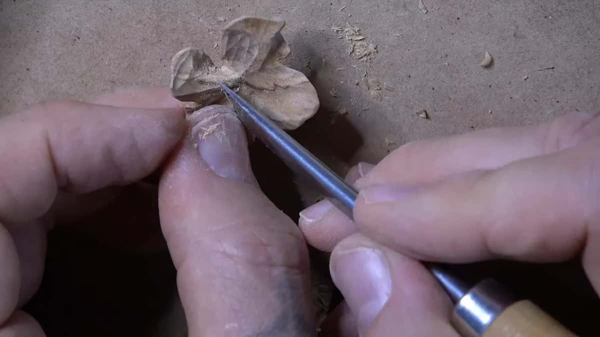 A narrow gouge shapes the petals of a small carved wood flower, shavings scattered across the bench.