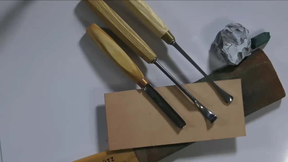 Three carving tools laid across a tan leather strop, with a grey compound lump and a green stick beside them.