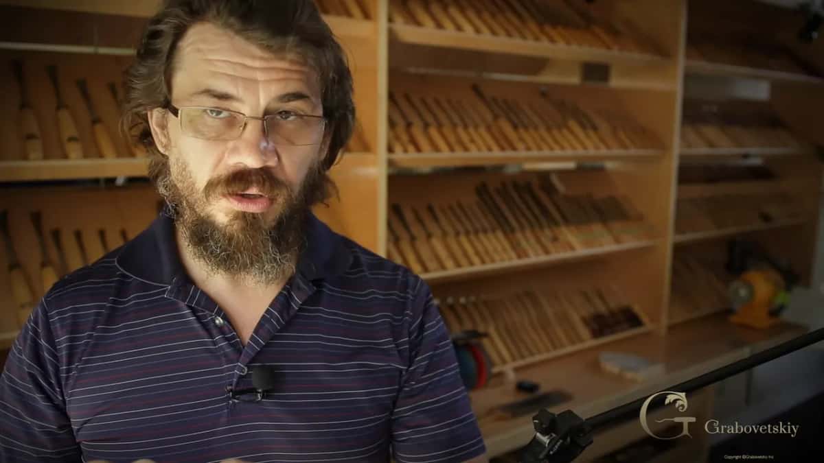 Rows of carving gouges fill the shelves behind a bearded man talking to the camera near a bench grinder.