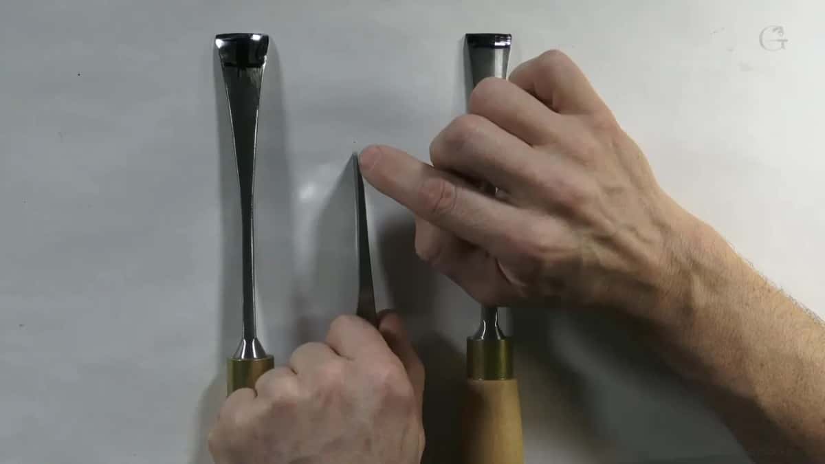 A finger touches the tip of a blade mounted between two wood-handled gouges on a plain gray wall.