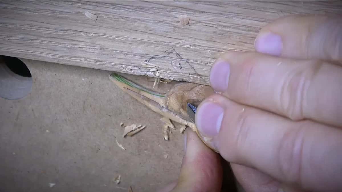 Thin wood tab at the corner of a board gets trimmed by a knife, fine shavings falling below.