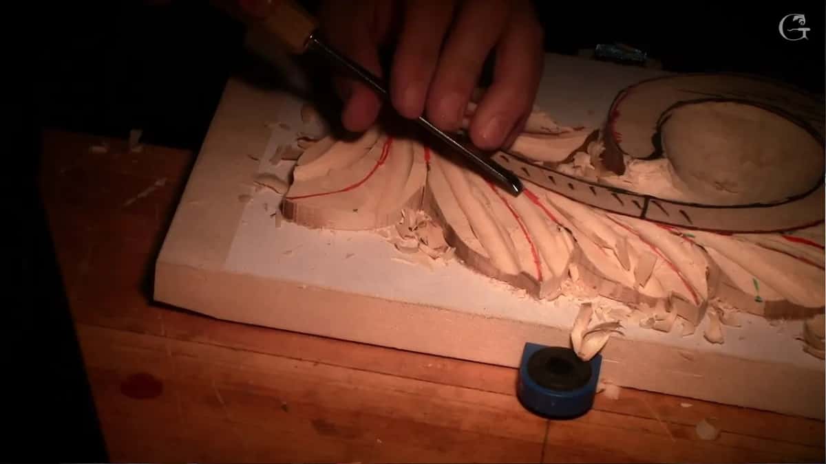 A small gouge lifts a pale curl of wood off a carved leaf; the curl stands on a roll of blue tape.