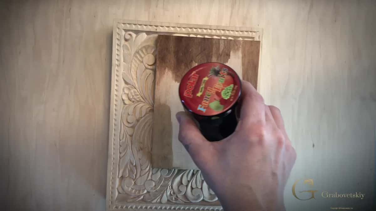 A hand holds a small dark jar above a test board half covered in brown stain on a pale carved panel.