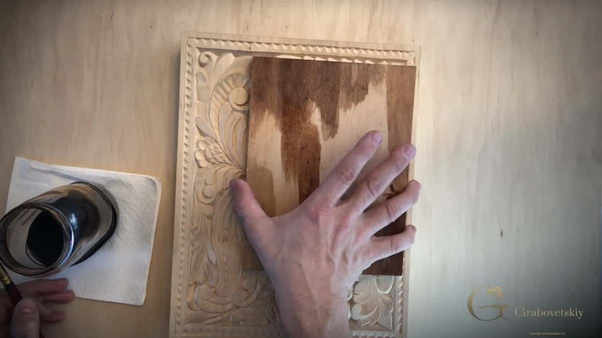Brown stain gets spread by hand across a board laid on a light carved panel, an open can waiting nearby.