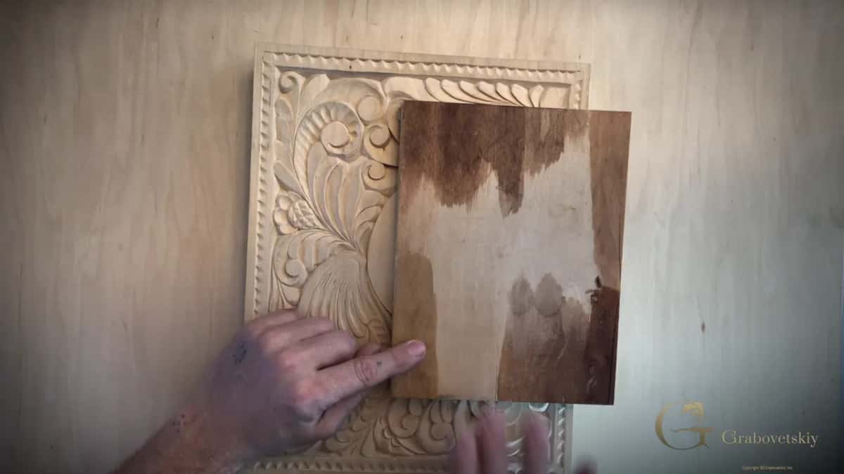 Half stained and half bare, a small board is held against a pale carved panel while a finger points at the line.