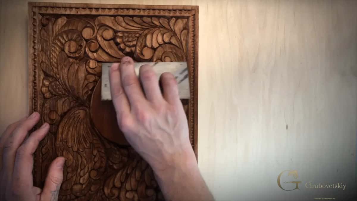 One hand presses a small flat block over the raised leaves of a deep brown carved panel.