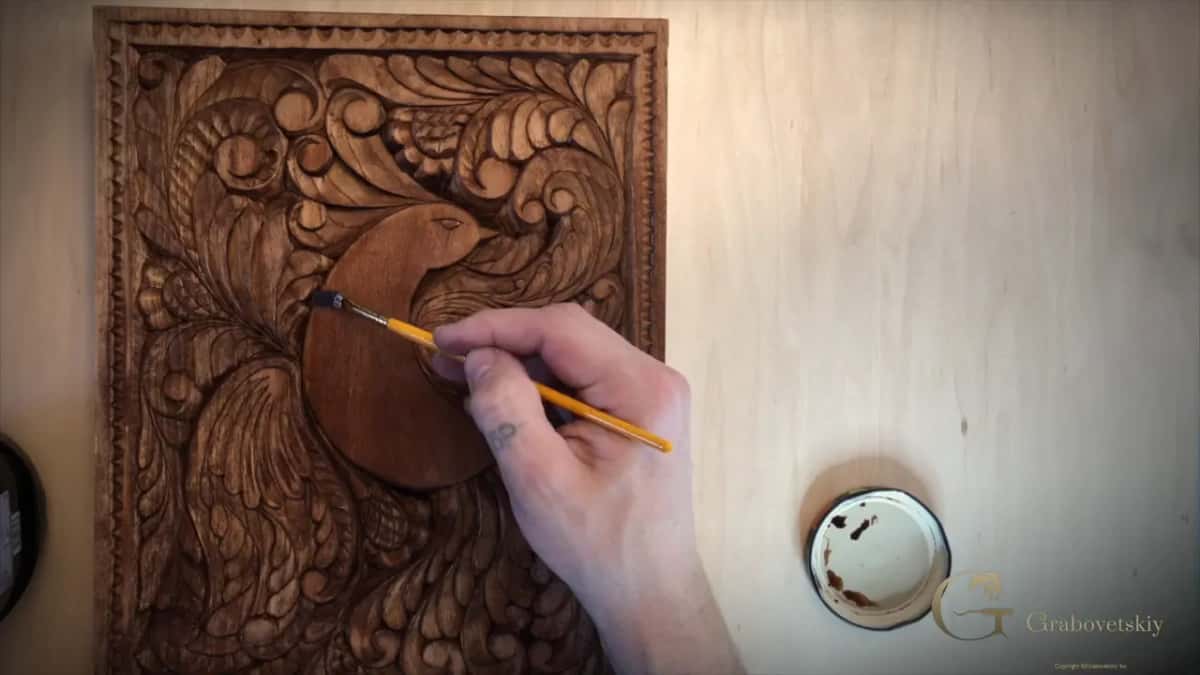 Dark finish goes onto the bird at the center of a rich brown carved panel, a small dish of liquid to the right.
