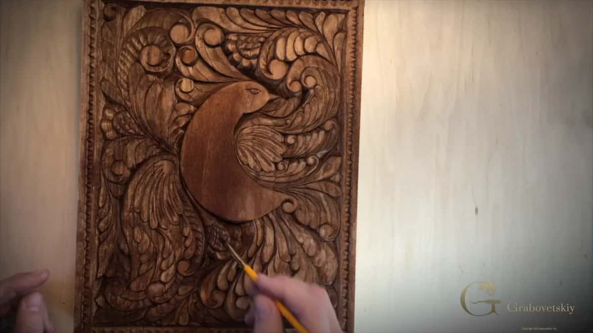 A brush touches the lower leaves of a deep brown carved panel, two hands steady at the bottom edge.