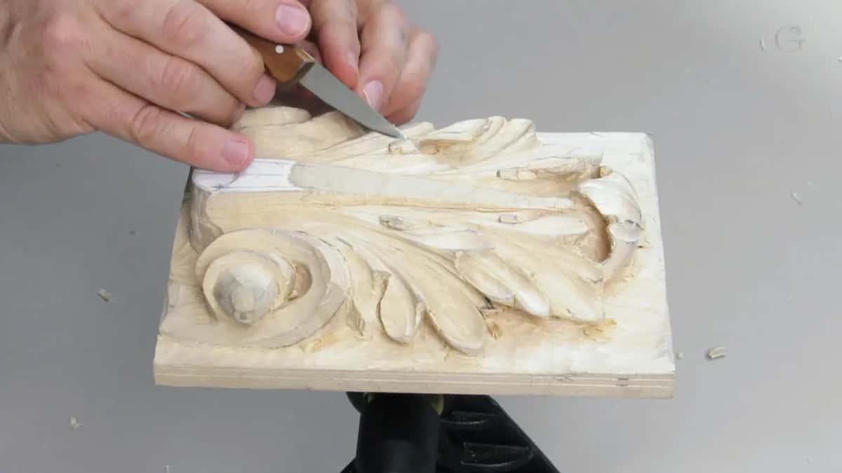 Guided by one hand, a small knife runs along a leaf on a pale carved block, chips lying on the grey bench.