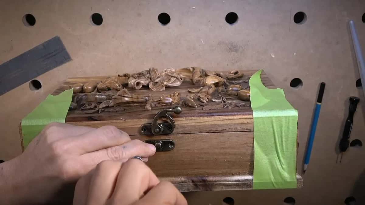 Hands set a brass catch into the front of a dark carved box, green tape masking both ends.