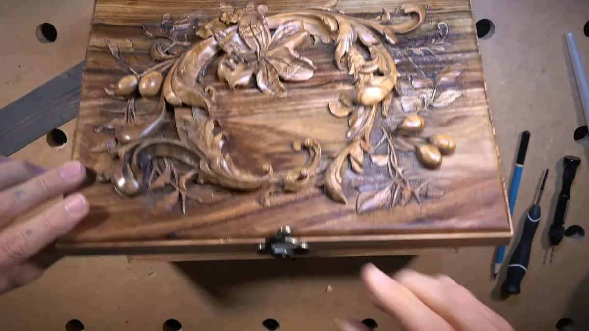 From above, a dark brown box lid shows a deep floral wreath, small tools resting on the bench beside it.