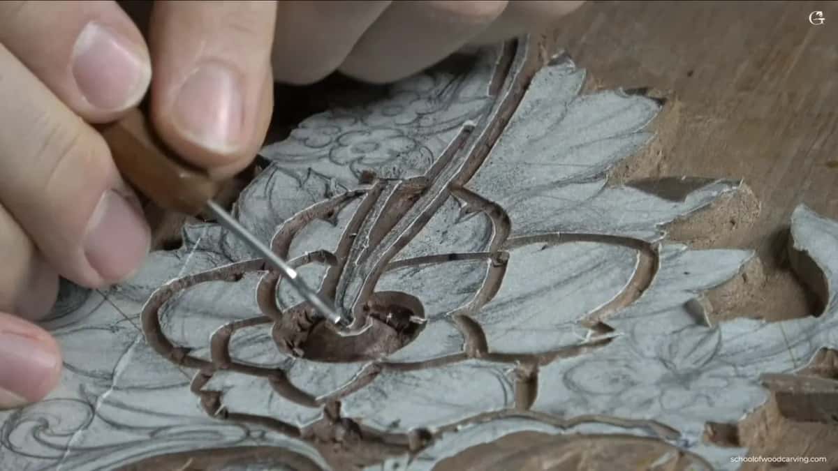 Fingers steady a slim tool at the middle of a white flower carved into a brown panel.