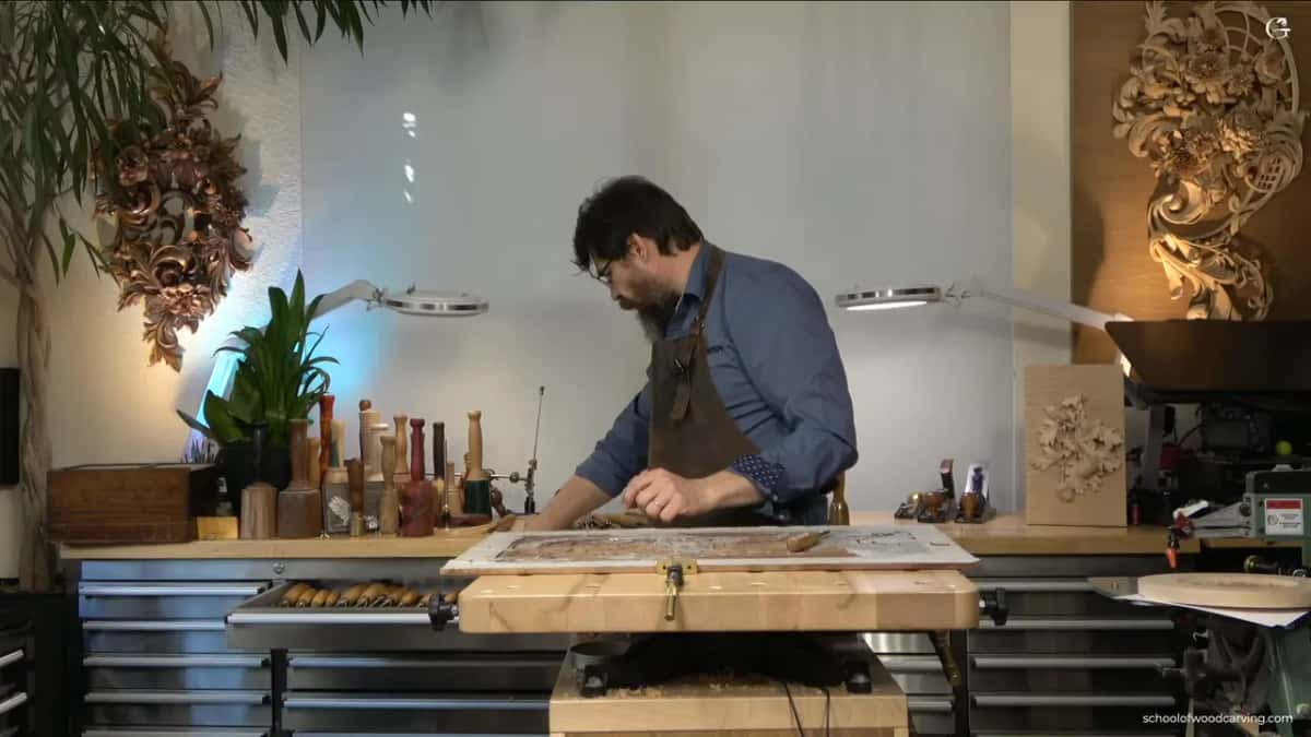 A carver leans over a long panel clamped to his bench, carved ornaments hanging on the studio wall behind.