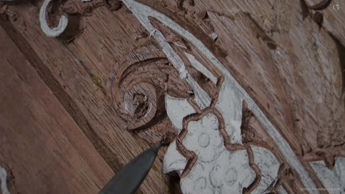 The point of a blade rests beside a carved scroll on brown wood scattered with small pale chips.