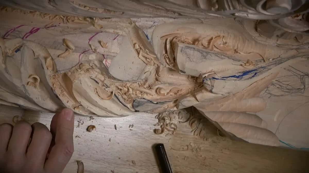 Fingertips tucked under a thin carved edge, shavings piled beneath it