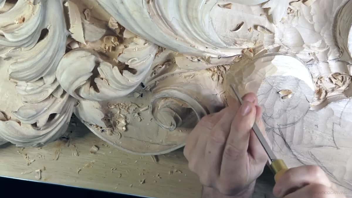 A slim tool goes into pale wood next to a carved scroll, both hands on it.