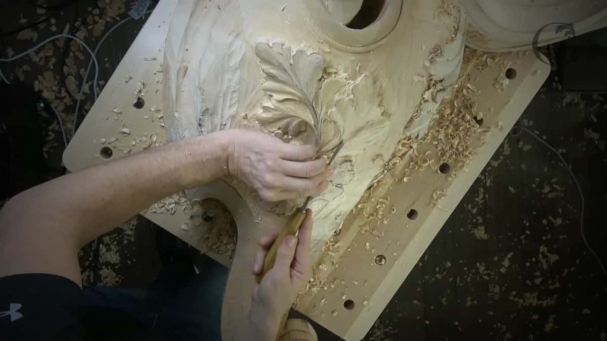 An arm reaches across the bench, and the hand below it holds a gouge low on the leaf.