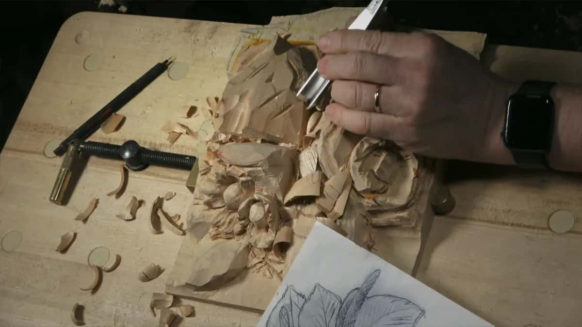 A hand holds the gouge low on the panel's top, the paper drawing below it