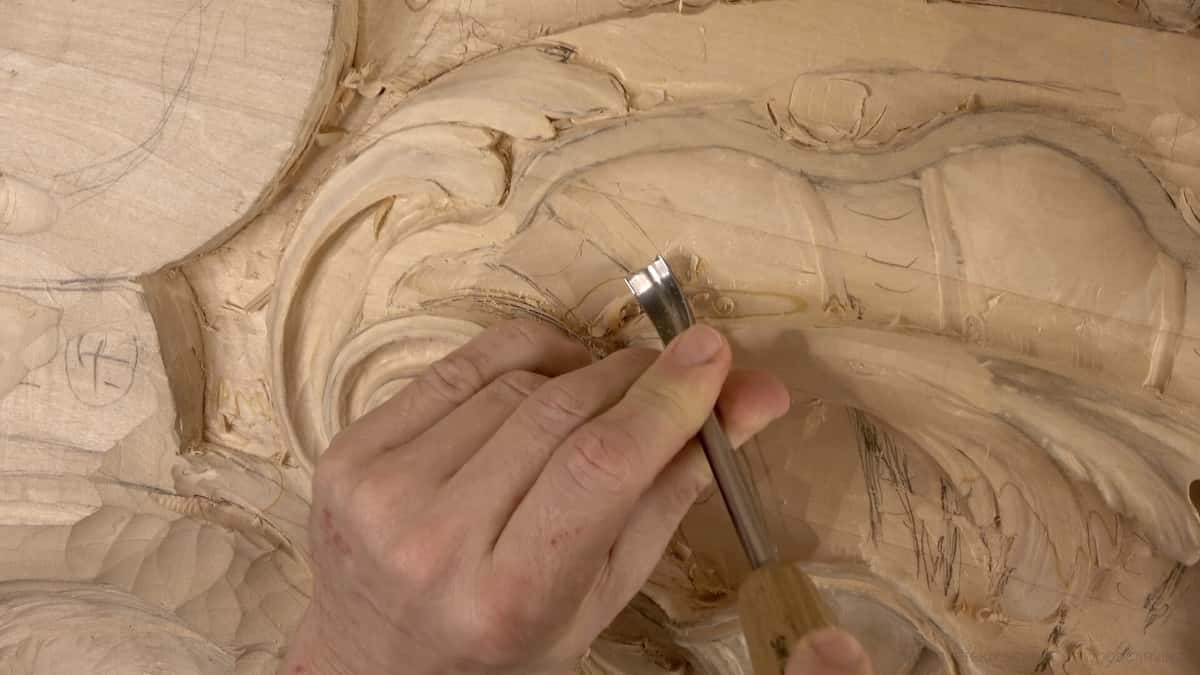 Overhead: a mallet and gouge in two hands on a panel drawn out in pencil, chips over the surface.