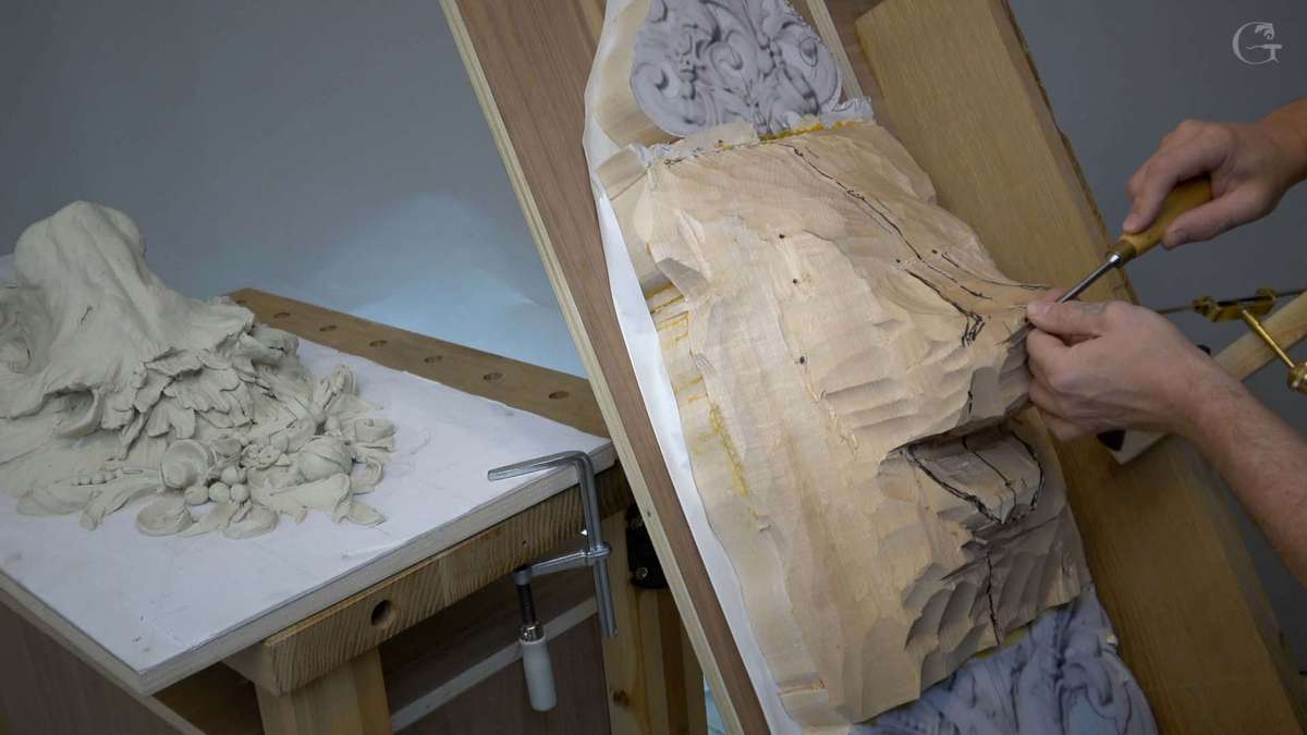 A person is focused on wood carving, skillfully using a chisel on a wooden piece that's clamped to a workbench. On the side, there's a clay sculpture of flowers and fruits, showcasing the creative process. The setting gives off a friendly, artistic vibe, perfect for wood carving classes and lessons.