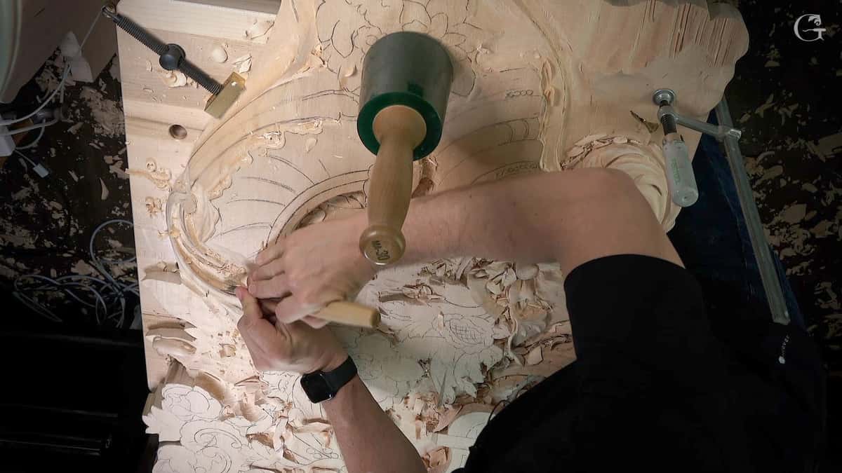 One hand guiding a bent gouge into a rococo shell, pencil layout lines across the wood around it.