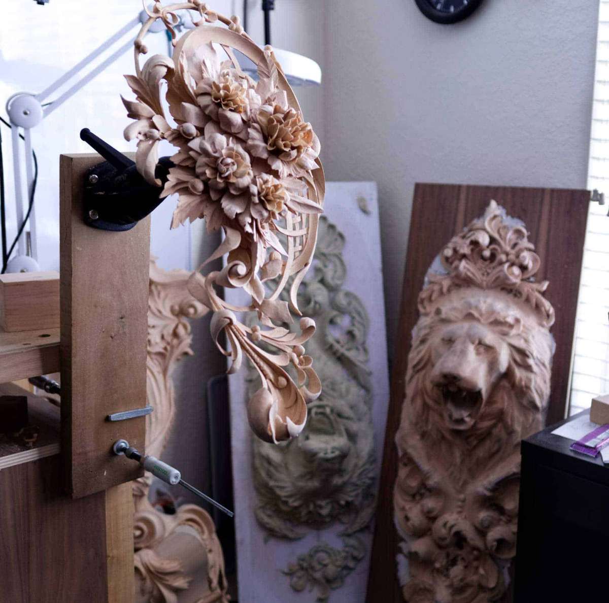 Alt text: A wood carving display showcasing intricate designs on a workbench. The foreground features a detailed floral piece crafted from light wood, while in the background, there are ornate lion carvings and other sculptures. This scene captures the essence of wood carving classes and lessons, emphasizing the artistic craftsmanship involved.