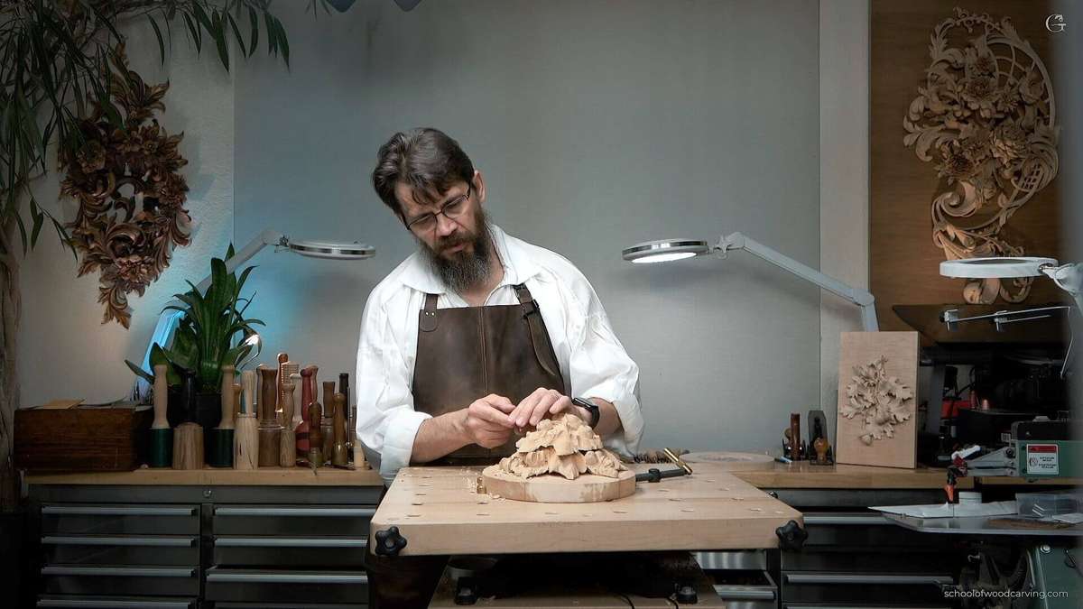 A woodcarver sits at a solid workbench holding carving tools, showing a proper woodcarving workbench setup