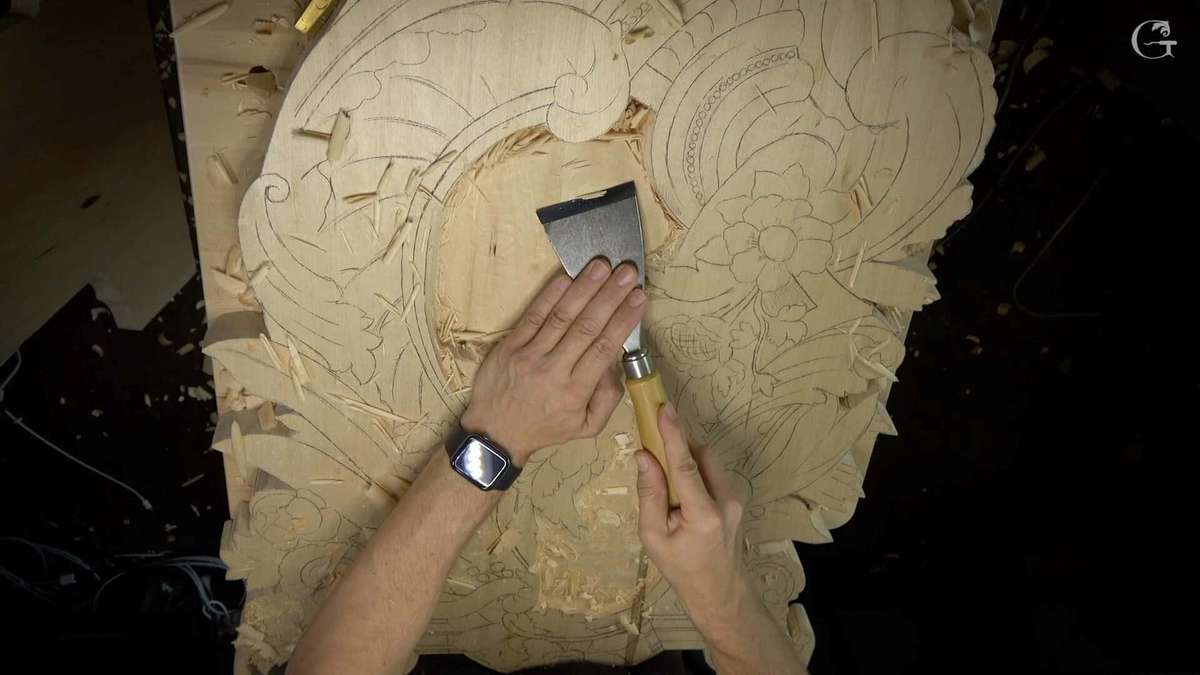 Close-up of hands using a chisel to carve intricate designs into a wooden board. The woodworker, wearing a watch on their left wrist, demonstrates techniques found in woodcarving tutorials online.