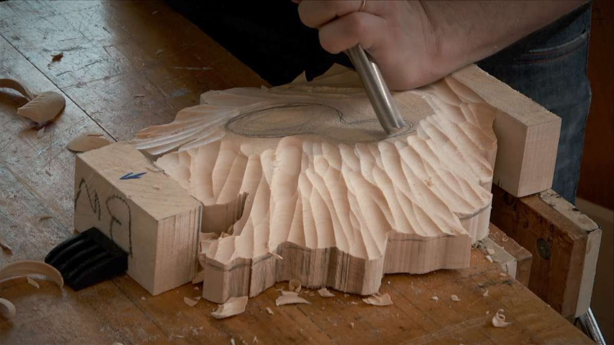 A person uses a chisel to carve a detailed pattern into a piece of wood on a workshop table, taking inspiration from woodcarving tutorials online.