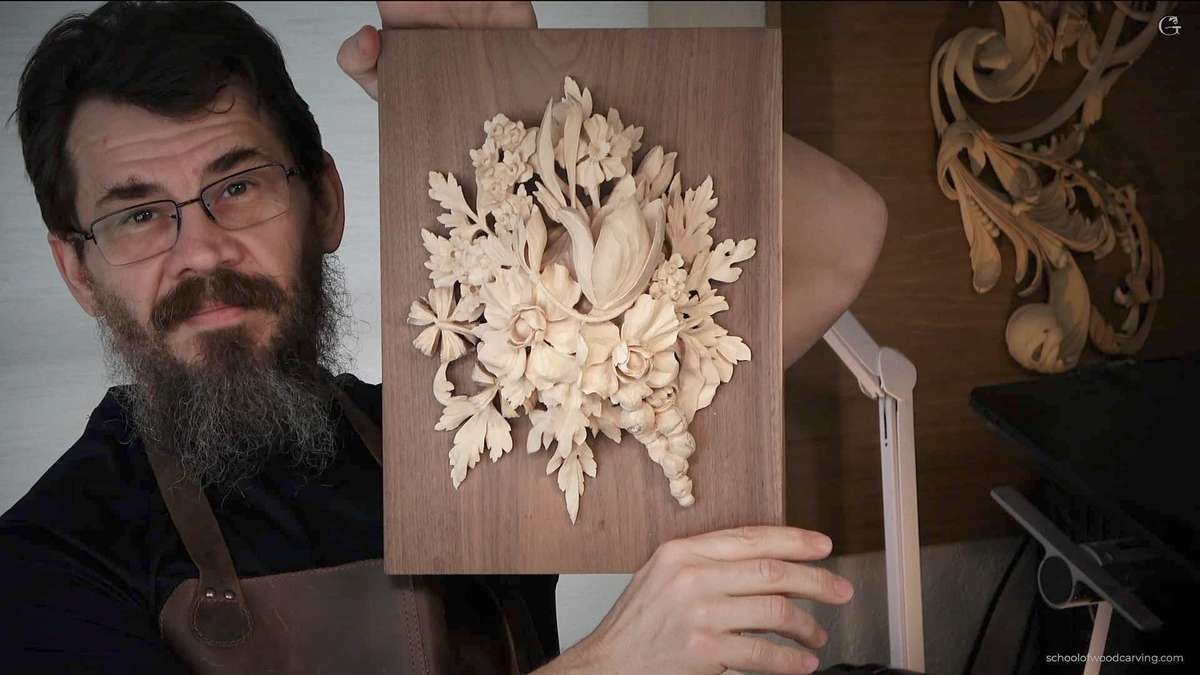 A man with glasses and a beard holds up a wood carving of flowers and leaves. He is wearing a leather apron, and the background includes more wooden carvings, reminiscent of the detailed projects you can learn in woodcarving tutorials online.