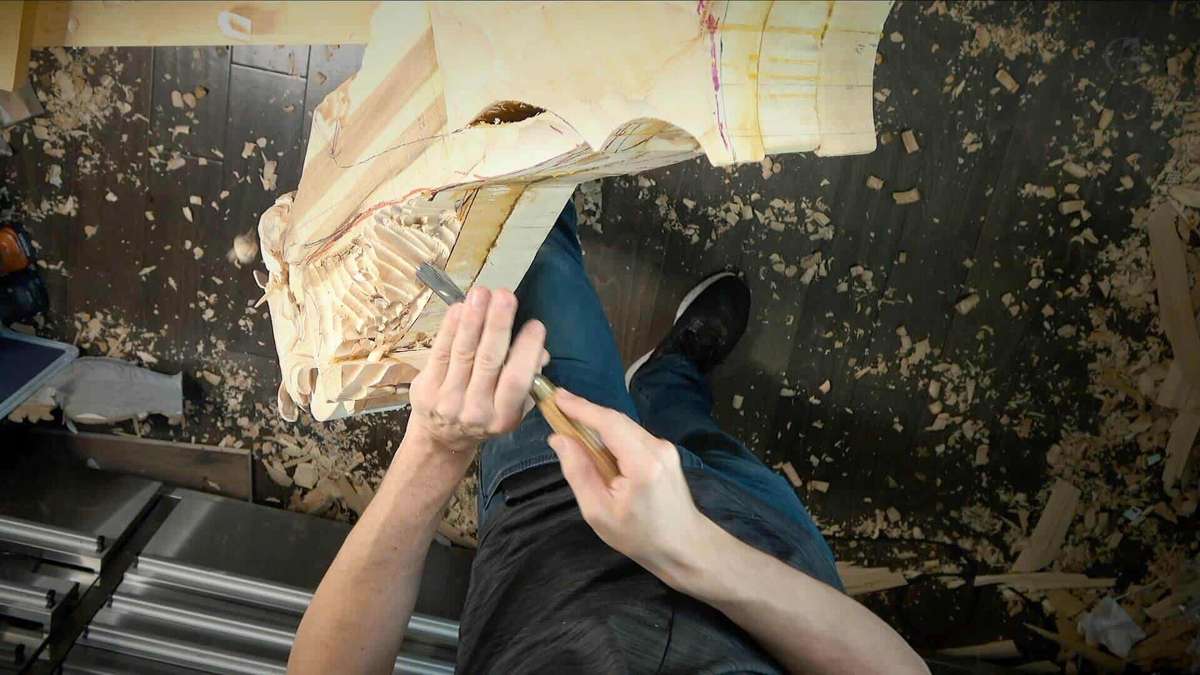 A person carves intricate details into a piece of wood with carving tools, surrounded by wood shavings on the floor. The image, taken from an overhead perspective, could easily be part of a detailed woodcarving tutorial online for enthusiasts.