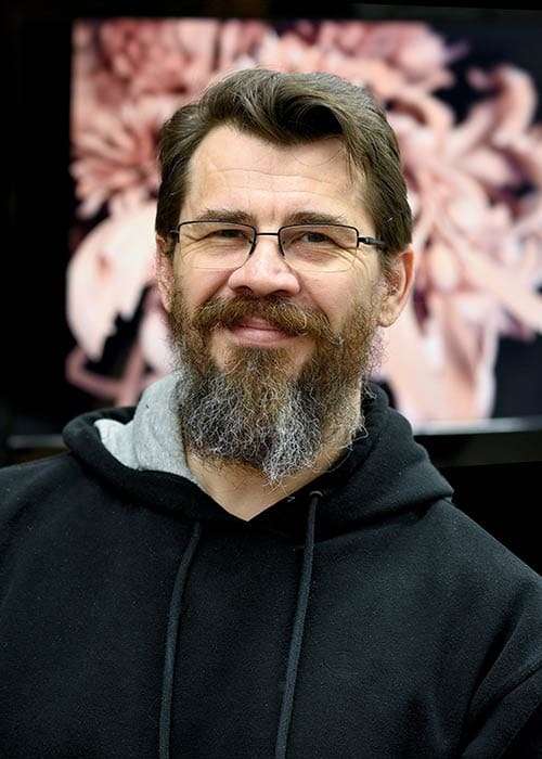 Smiling person wearing glasses and a hoodie, with a full beard. In the background, there's a blurred image of flowers. This could be someone involved in wood carving, possibly offering wood carving classes or lessons.