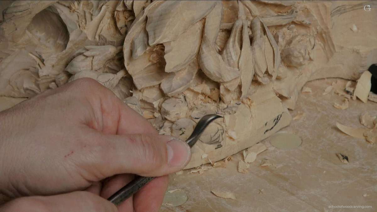 WOODCARVING LESSONS: A close-up photo shows Woodcarver Grabovetskiy holding a carving tool. He carefully carves detailed flower and leaf shapes into wood. You can see wood shavings and carved patterns on the wood's surface. The table has small pieces of wood scattered around. Learn how to carve beautiful designs with expert Woodcarver Grabovetskiy in our easy-to-follow woodcarving lessons or course. See the tools, clear carving steps, and tips for beginners in every tutorial picture. - Wood Carving tutorials