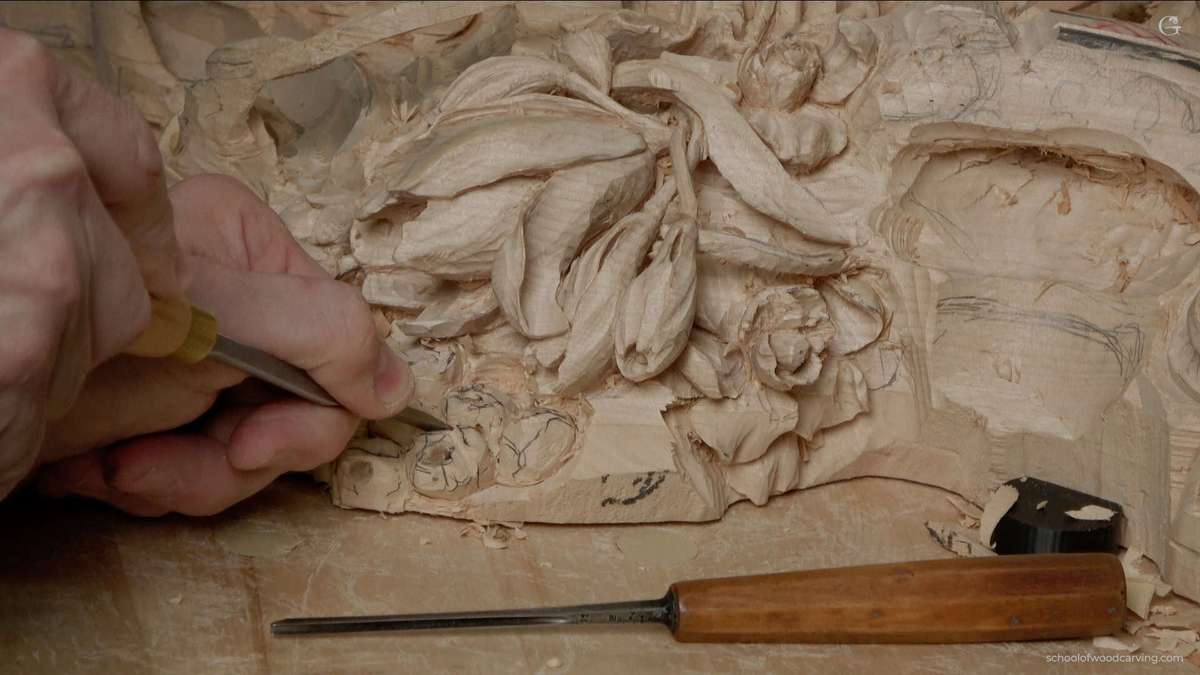 WOODCARVING LESSONS: Close-up photo of Woodcarver Grabovetskiy’s hand using a chisel for woodcarving. He is carving beautiful flower patterns into light wood. Another chisel is on the table next to the wood. This picture shows detailed and careful woodcarving work during a lesson or tutorial with master Grabovetskiy. Learn how to carve floral designs in wood step by step in this course. - Wood Carving tutorials