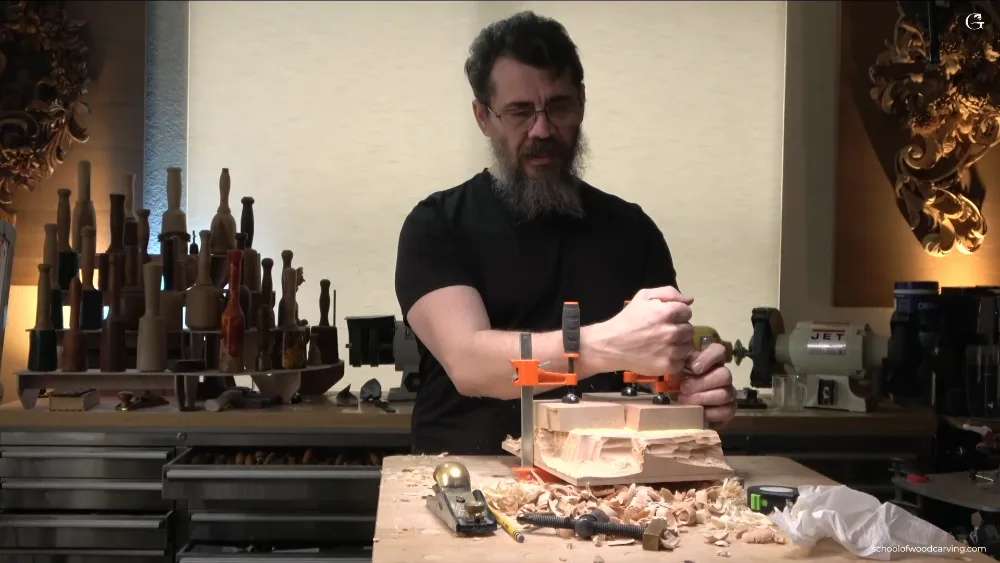 WOODCARVING LESSONS: Woodcarver Grabovetskiy clamps wood at a workbench with tools and shavings. Hazelnut carvings and spindles shown behind him. - Wood Carving tutorials