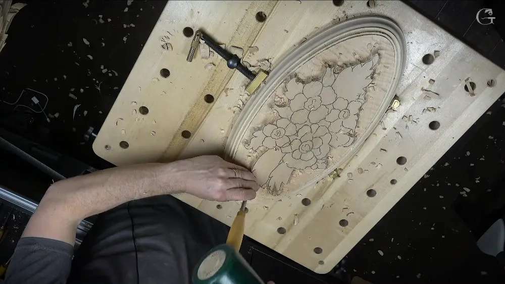 A carver's hand guides a cutting tool across a relief carving board filled with flower designs
