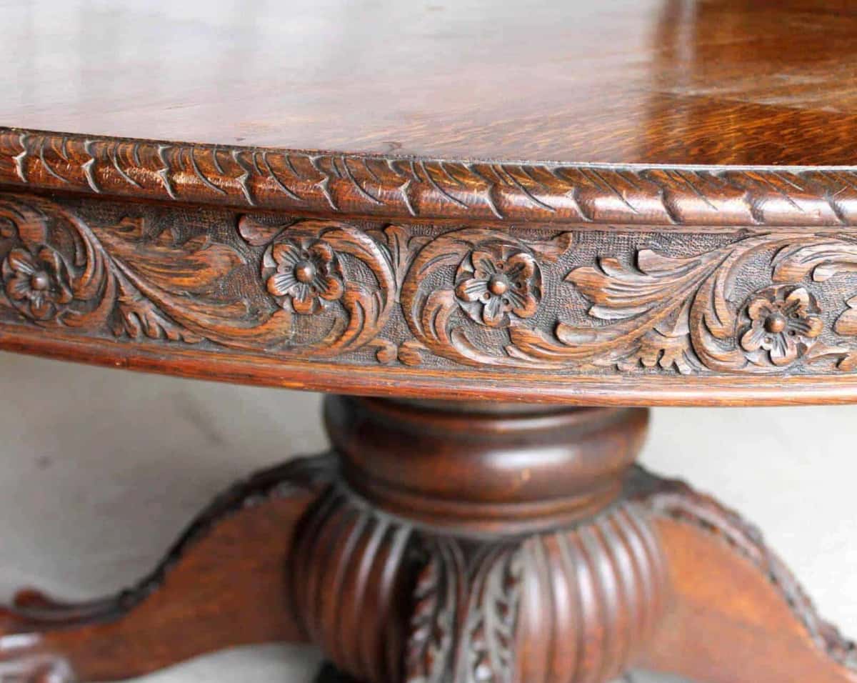 Antique oak dining table close up, its carved apron dark with age and rubbed bright along every raised petal