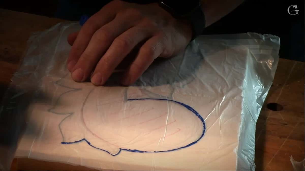 Tracing the template in blue Blue marker outline traced onto a sheet of plastic laid over a drawing, one hand steadying the sheet flat
