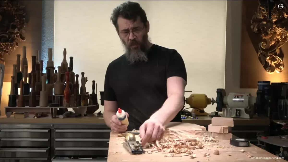 Glue bottle in one hand at the bench, shavings and a small brass plane on the top, carved panels lit on the wall behind