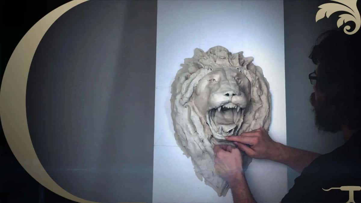 Clay lion head roars off a vertical board while both hands press the mane into shape under a work light