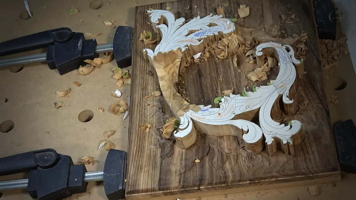 A white paper pattern of a scroll glued to a clamped dark board, pale carved wood showing through, chips all over.