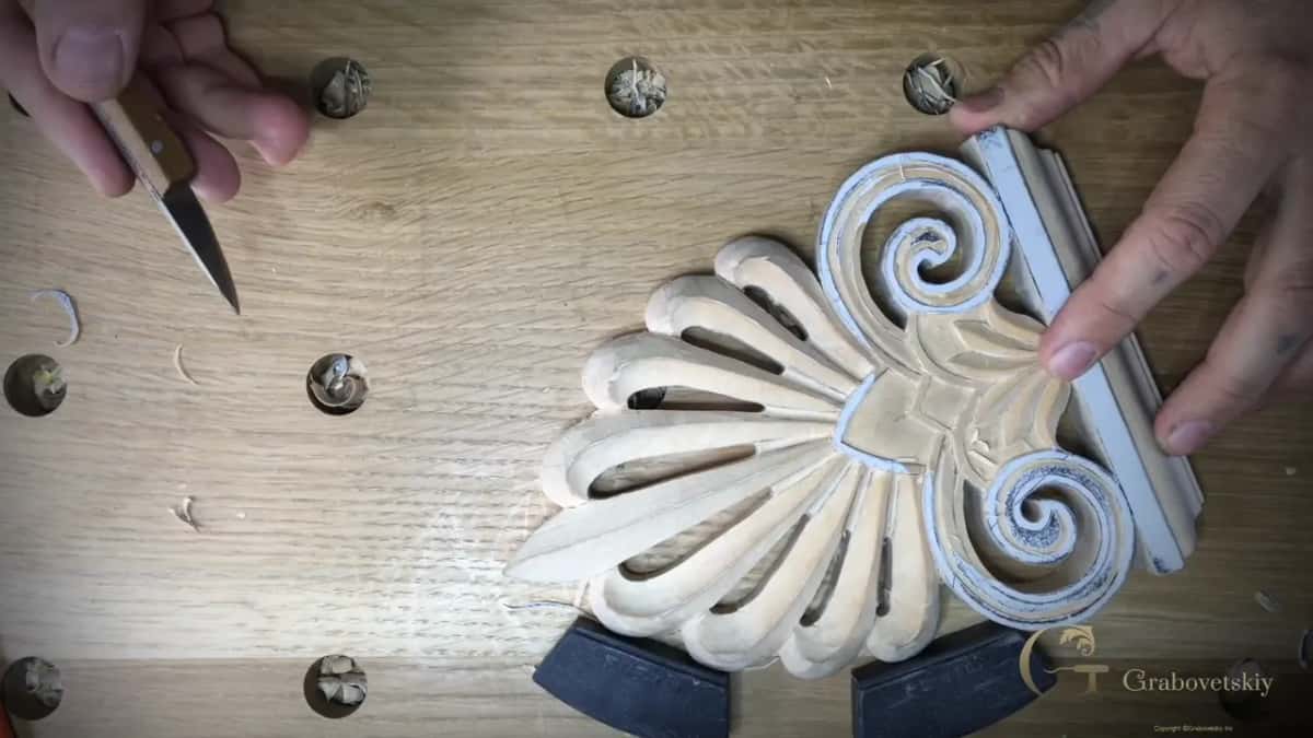 Straightedge held against a carved fan palmette with chalked spiral volutes, a carving knife in the other hand