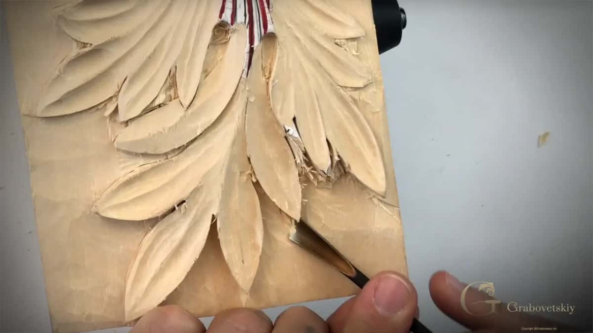 Bent gouge reaching under a carved acanthus lobe, fingers guiding the blade from below the panel