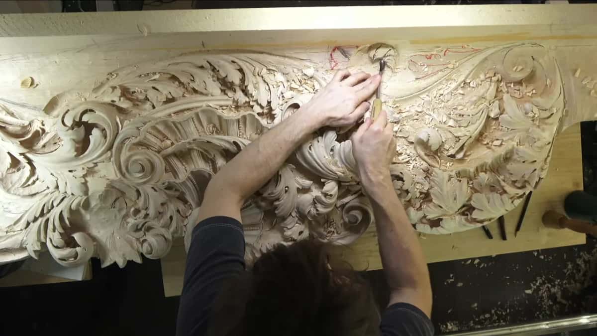 Seen from above his shoulder: both hands drive a gouge into a long acanthus frieze buried in chips