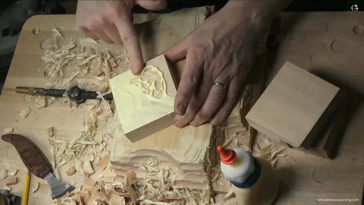 Yellow wood glue spread in a swirl across the back of a small carving blank while both hands wiggle it down onto the backing block, a spare block and a knife waiting on the bench