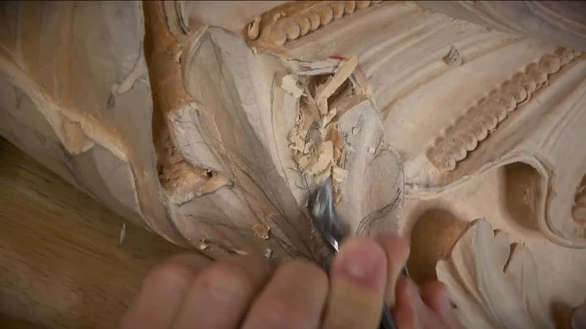 A gouge tip lifts a ragged tuft of fibre out of a carved leaf, pencil layout lines still on the wood.