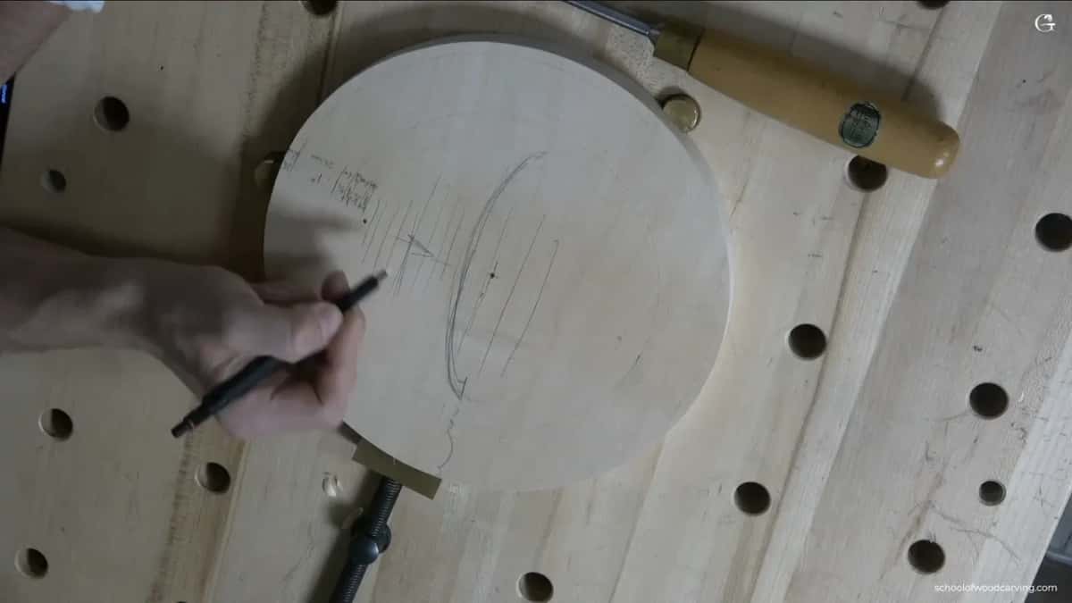 A round basswood blank clamped to a dog hole bench, first pencil lines going down, one gouge beside it.